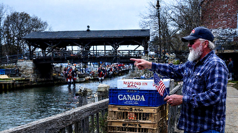 dock worker in weathered flannel shirt pointing at a crate of fish labeled "Made in Canada" while holding an American flag