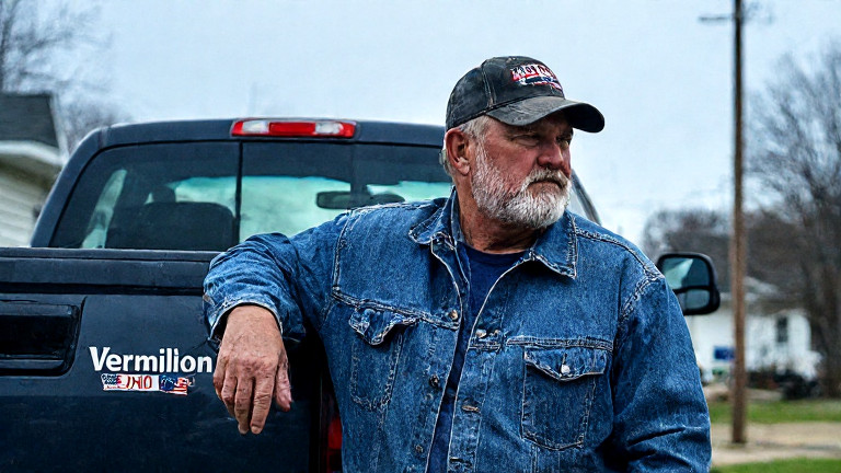 truck driver in worn denim jacket leaning against a pickup truck with "VERMILION, OHIO" bumper sticker, wiping sweat from brow