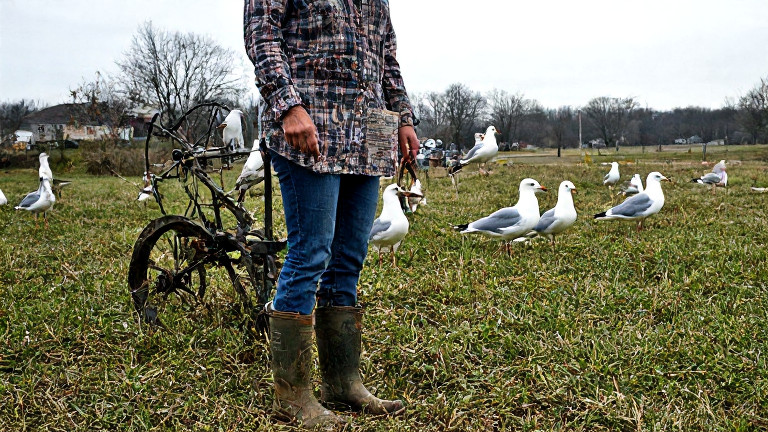 woman farmer in muddy boots leaning on a tractor while holding a Constitution-themed weather vane