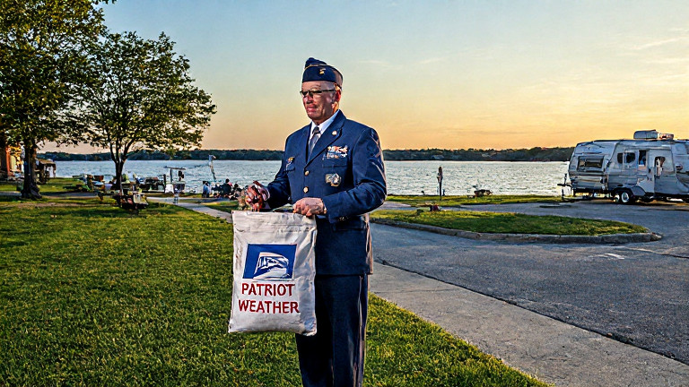 postal worker in vintage USPS uniform adjusting a "Patriot Weather" mailbag with a thermometer on a Vermilion street corner