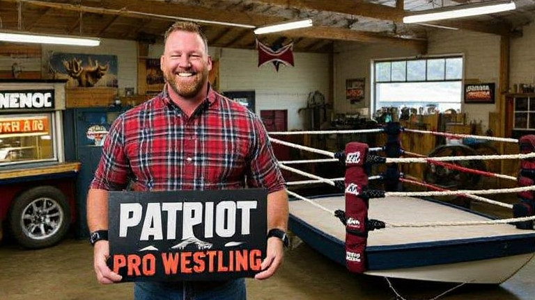 Vermilion wrestling promoter in plaid shirt holding a "Patriot Pro Wrestling" sign beside a vintage wrestling ring in a cluttered community center