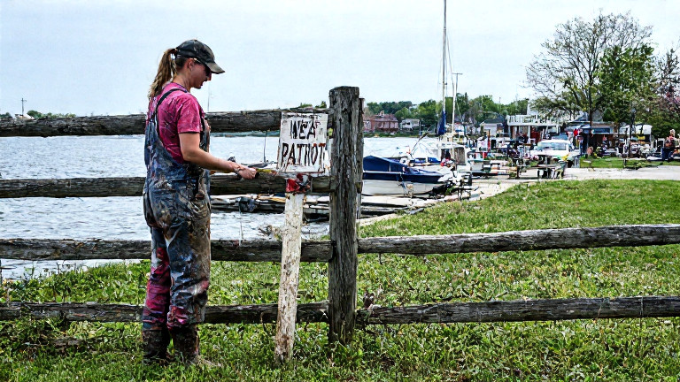 woman farmer in mud-splattered coveralls inspecting a weathered "Patriot" sign nailed to a wooden fence post