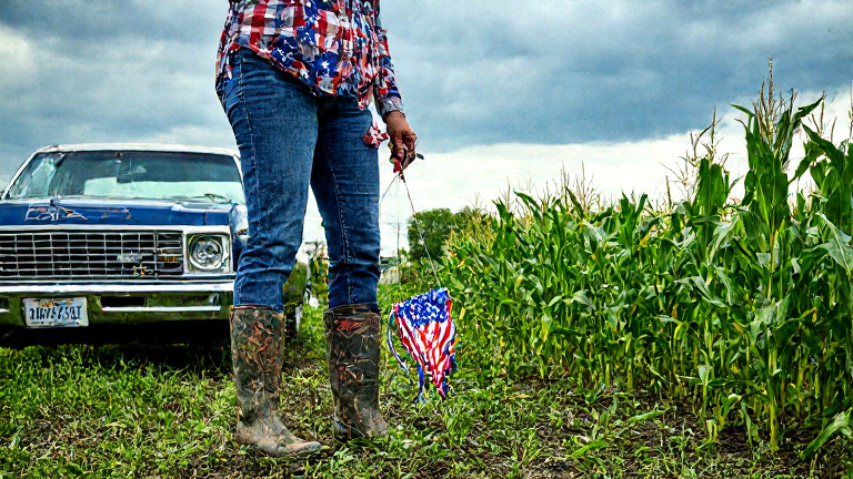 woman farmer in muddy boots holding a flag-patterned lure near a cornfield