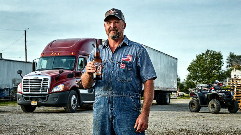 mechanic in grease-stained coveralls holding up a bottle of Liberty Lager next to a truck with a small American flag taped to the cab