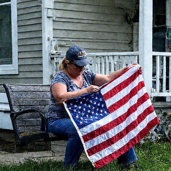 woman in faded military cap carefully stitching a large American flag on a porch swing