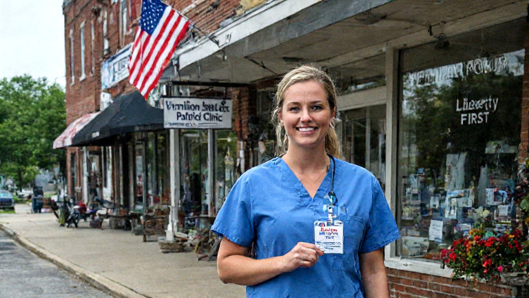 young nurse in scrubs holding a "Vermilion Patriot Clinic" badge while walking past a "Liberty First" banner in a downtown Vermilion storefront