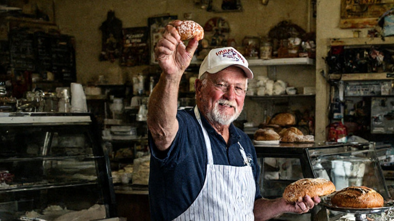 woman baker in flour-dusted apron holding up a "Patriot Sausage" loaf at a local bakery counter