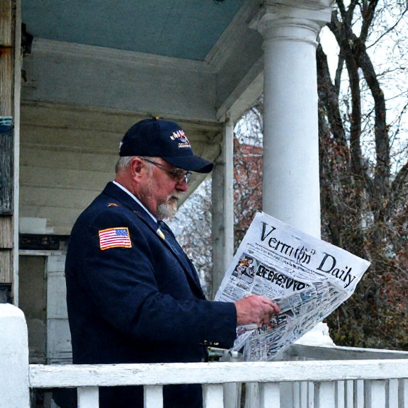 postal worker in uniform sorting mail on a porch with a "Vermilion Patriot Daily" newspaper visible