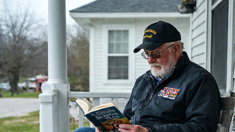 Vermilion veteran in Marine Corps jacket reading a book titled "Dolly: A Vermilion Hero" on a porch