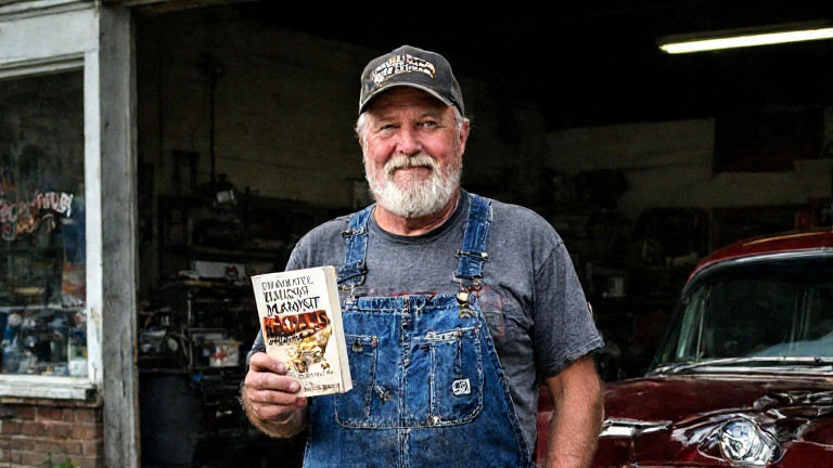 mechanic in worn coveralls holding a "Midnight Maas" book while leaning on a vintage car in a Vermilion garage