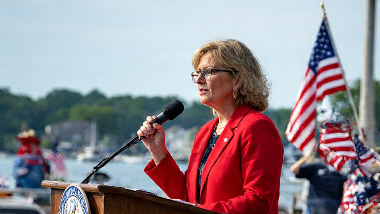Vermilion City Council member Linda "Patriot" Jenkins in a bright red blazer holding a flag, addressing a crowd at the Vermilion Waterfront Park