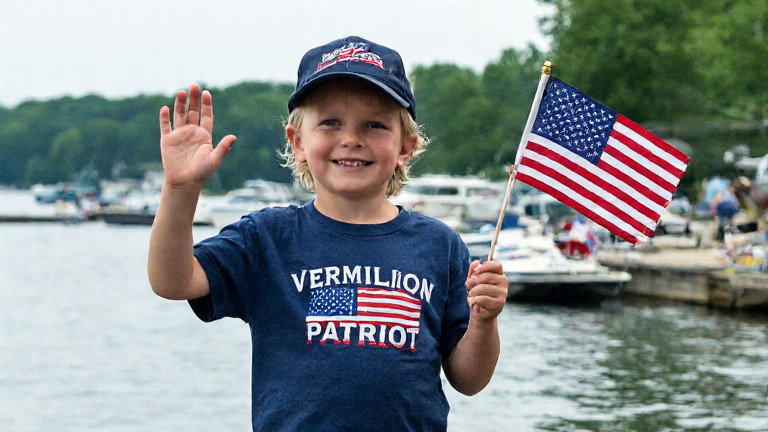 Older woman in a "Vermilion Patriot" t-shirt waving a small American flag in front of Lake Erie, smiling at boats